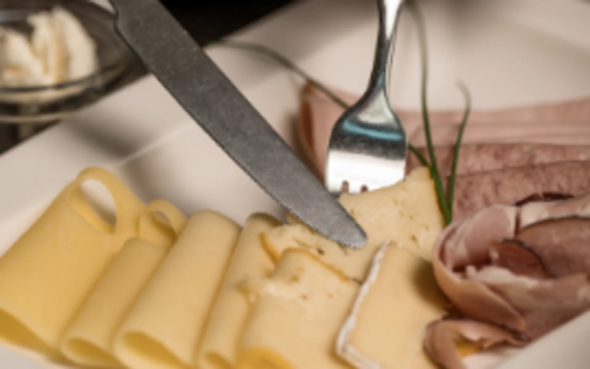 Person schneidet Käse auf Teller mit Wurst und Brot im Hintergrund