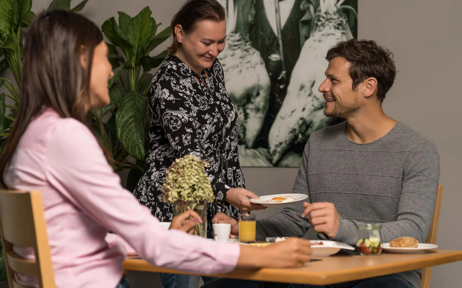 Frau serviert Mann ein Frühstücksei an einem Tisch mit einer weiteren Frau