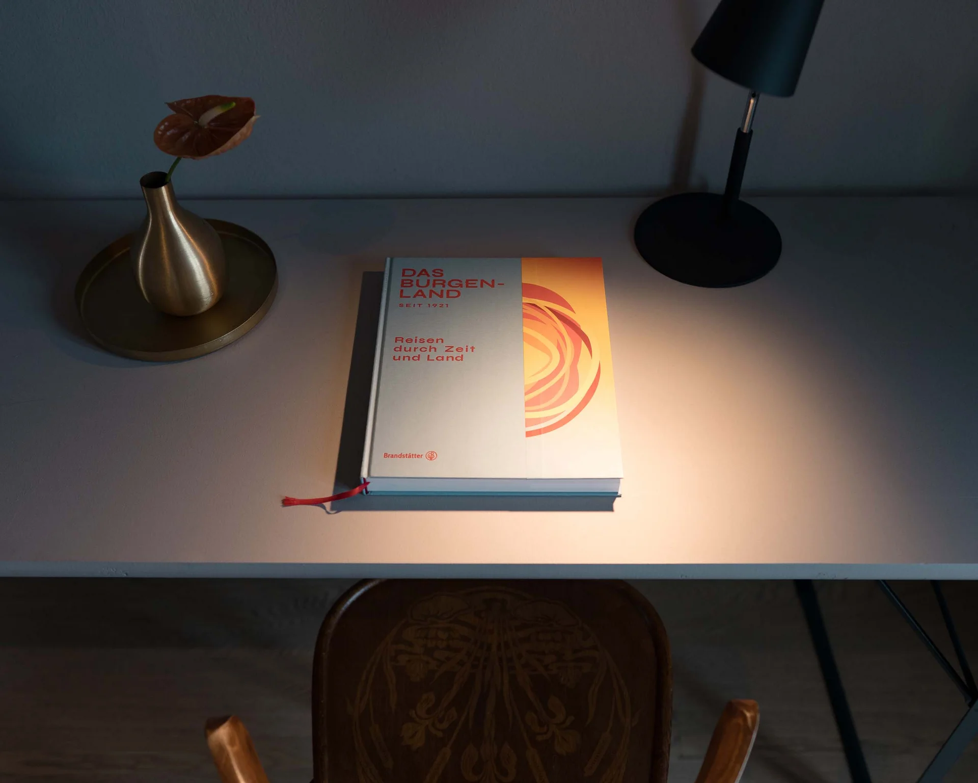 Schreibtisch mit Buch, einer kleinen Lampe und einer Vase mit Pflanze, darüber ein Stuhl